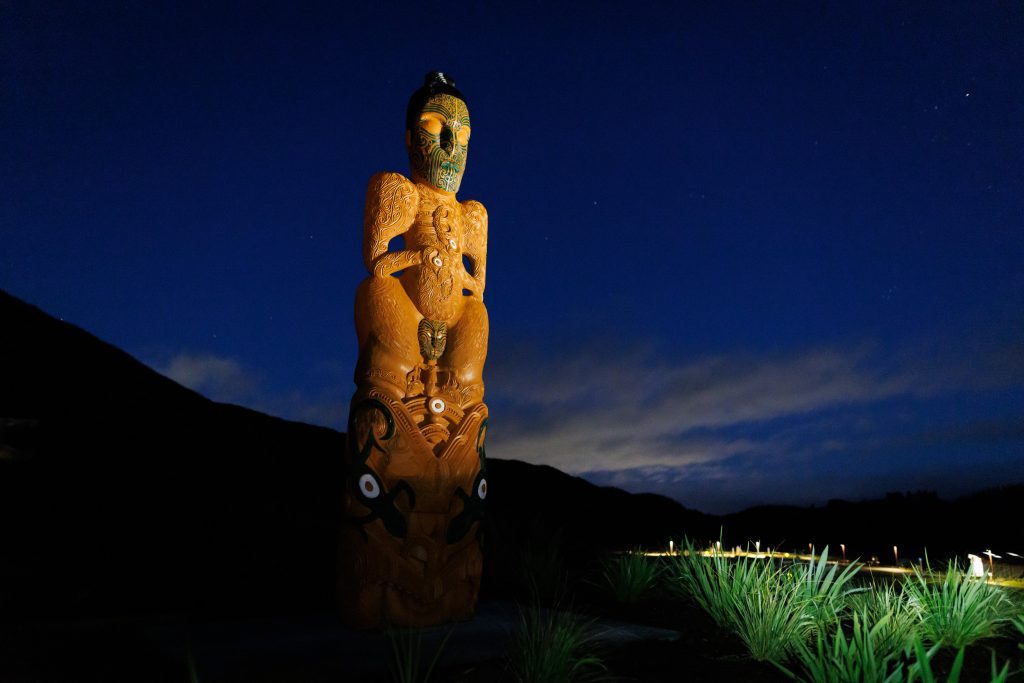 A māori sculpture of an umukaria (chief) on a hilltop at night lit up by lights with grass in the foreground